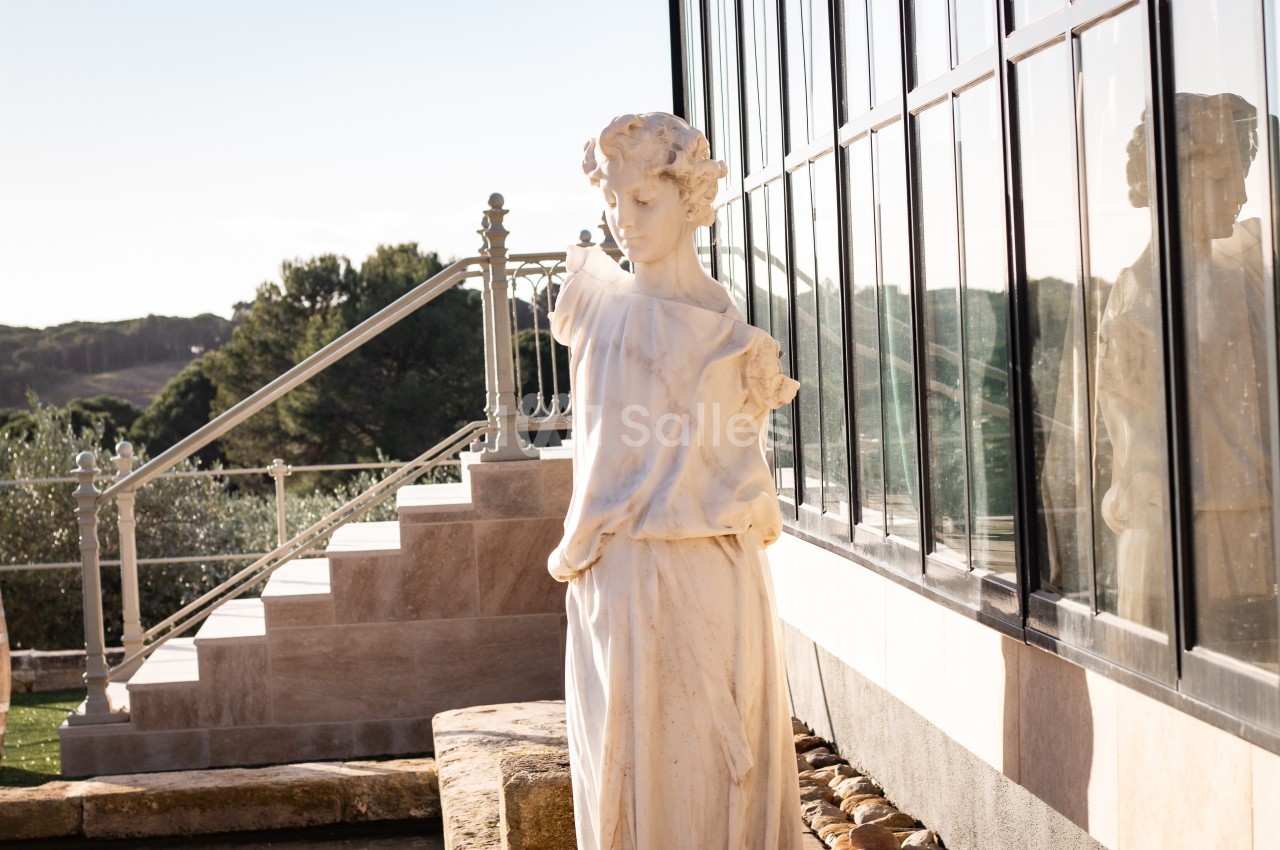 Statue en marbre représentant une femme, placée près d'une verrière avec un escalier et un paysage naturel en arrière-plan.