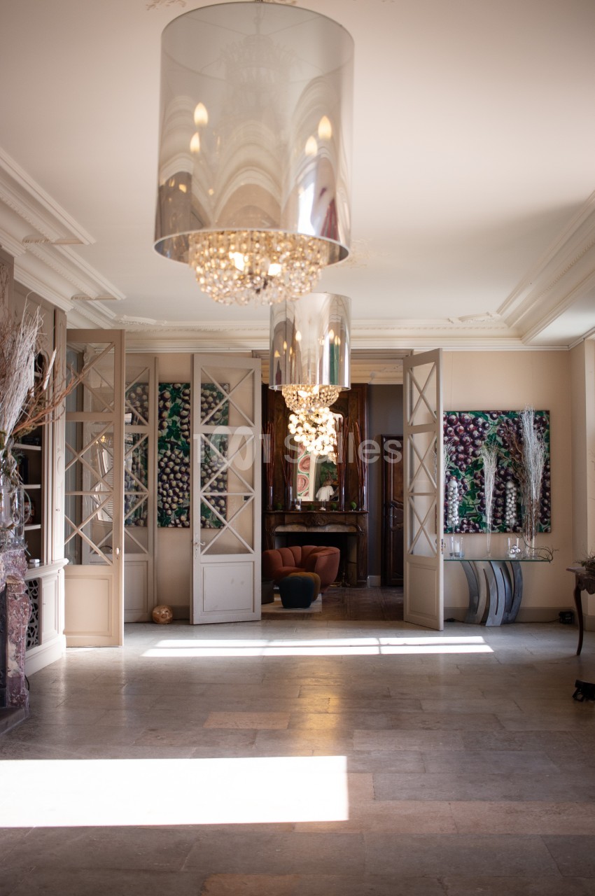 Intérieur lumineux avec lustres modernes, portes vitrées ouvertes et fauteuil visible dans une pièce adjacente.