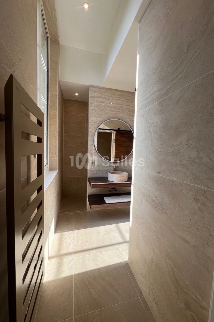 Salle de bain lumineuse avec murs en carrelage beige, miroir rond, vasque sur meuble en bois et sèche-serviettes.