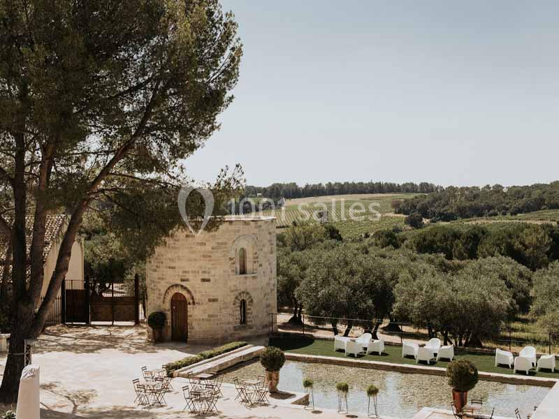 Cour ensoleillée avec bassin, chaises et tables, entourée d'arbres, d'un bâtiment en pierre et de vignes en arrière-plan.