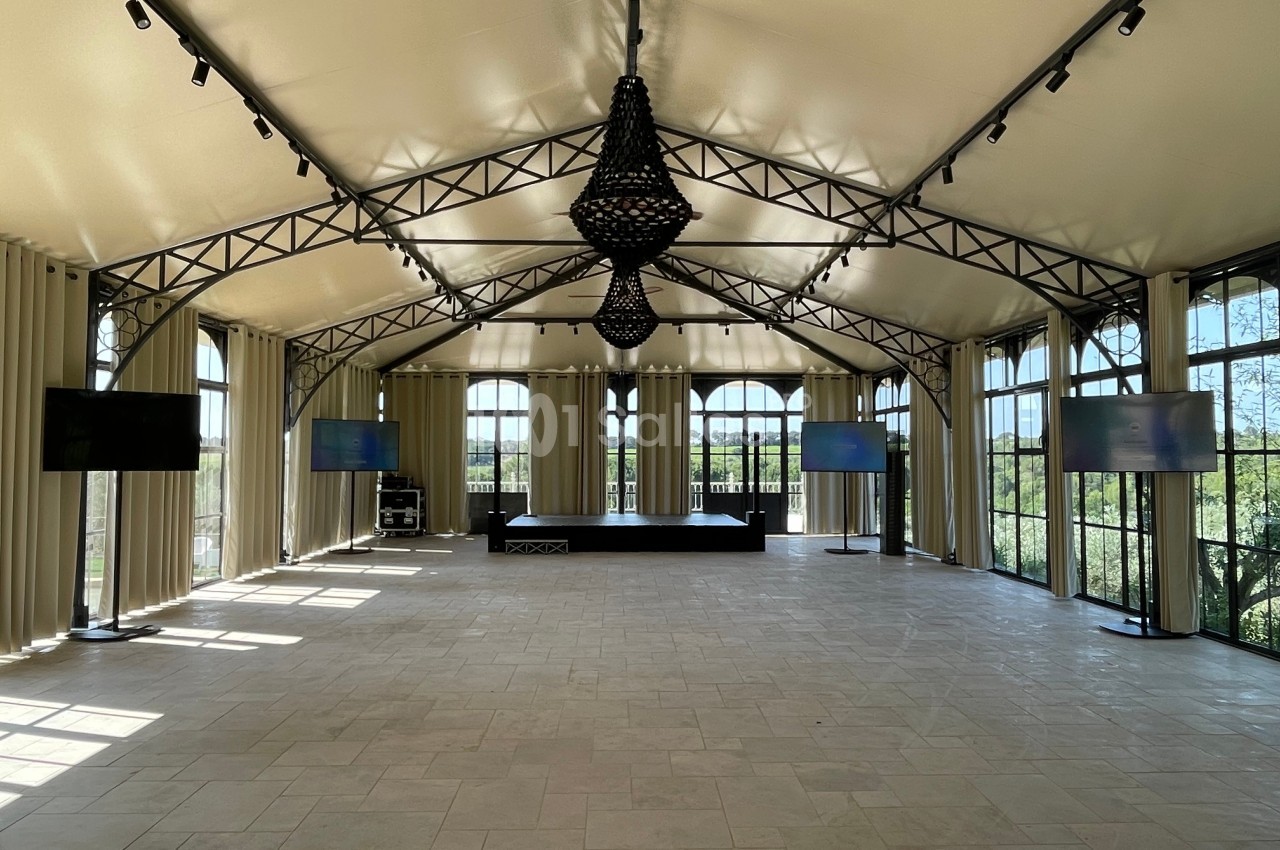 Salle lumineuse avec toit en toile, grandes baies vitrées, scène centrale et écrans disposés sur les côtés.
