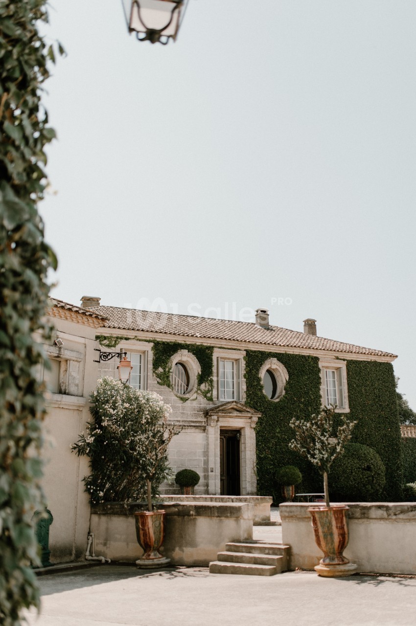 Façade d'une maison en pierre recouverte de lierre, avec des fenêtres rondes et des plantes en pots devant l'entrée.