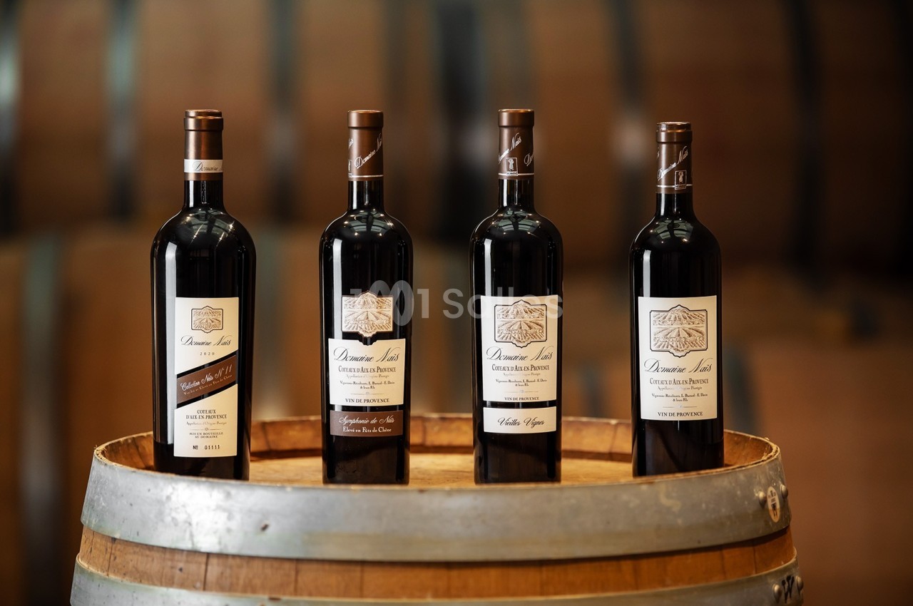 Quatre bouteilles de vin rouge alignées sur un tonneau en bois, avec des fûts flous en arrière-plan.