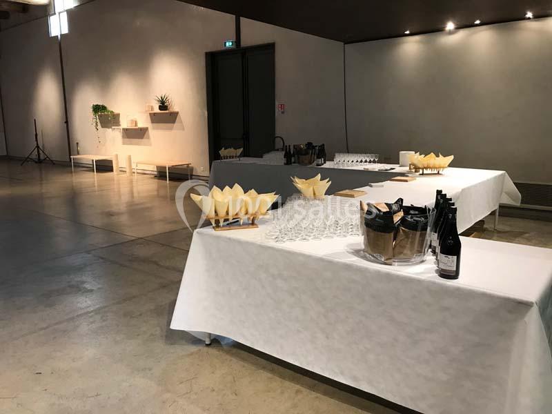Tables dressées avec des verres, bouteilles et amuse-bouches dans une salle sobrement décorée et bien éclairée.