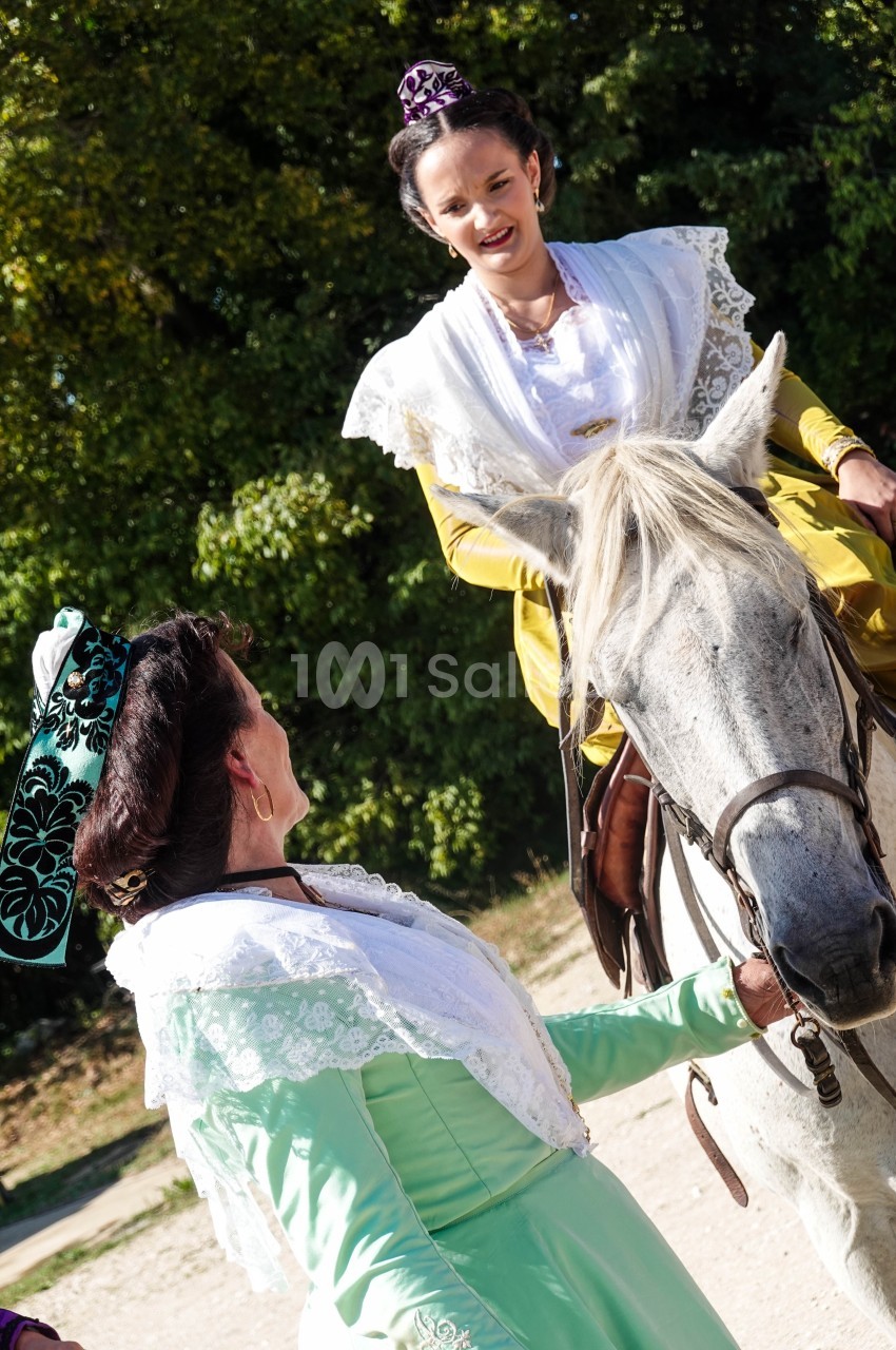 Deux femmes en tenue traditionnelle, l'une à cheval et l'autre à pied, discutent dans un cadre extérieur verdoyant.