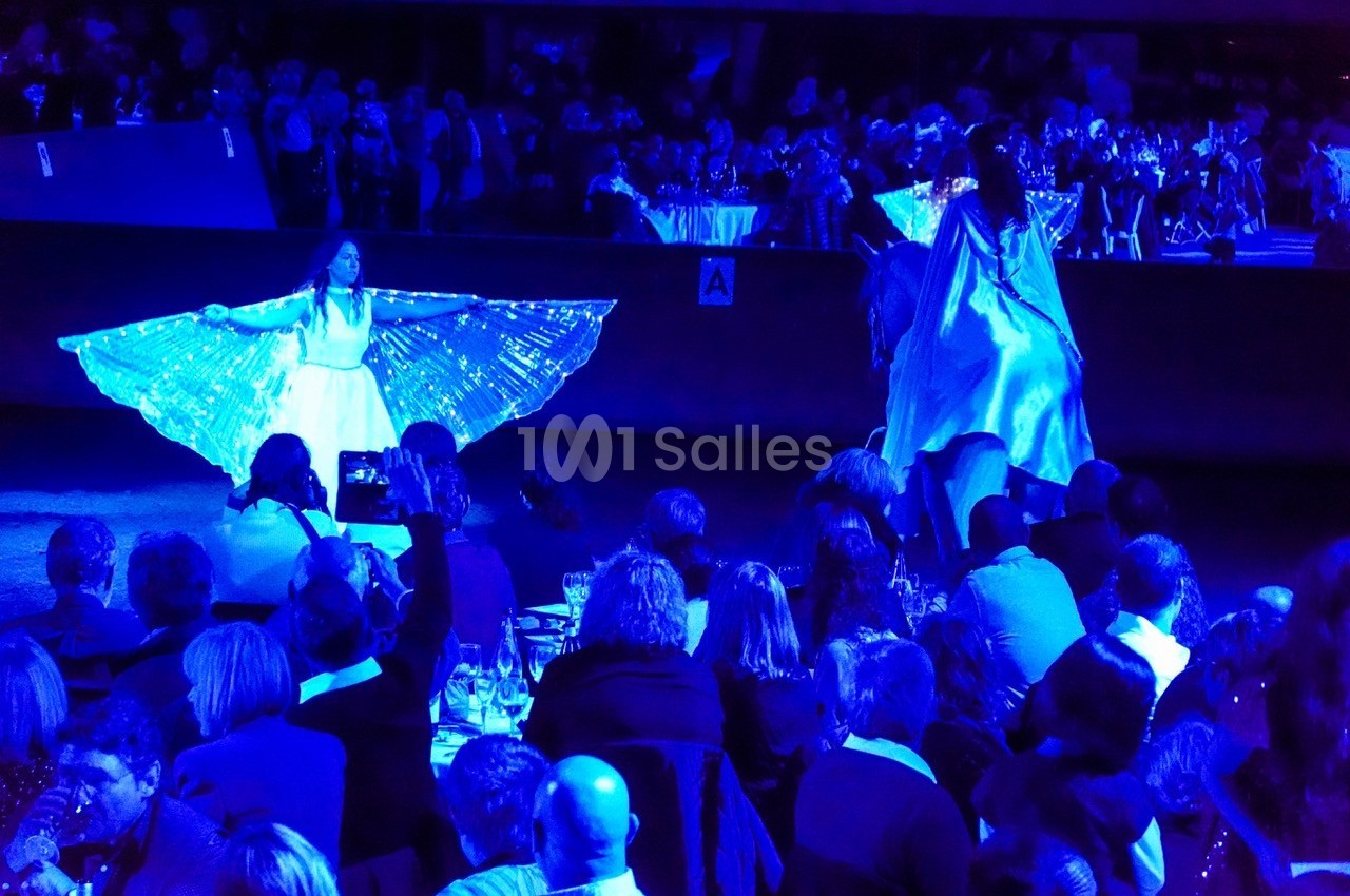 Des artistes en robes lumineuses dansent sur scène devant un public assis dans une ambiance tamisée bleutée.