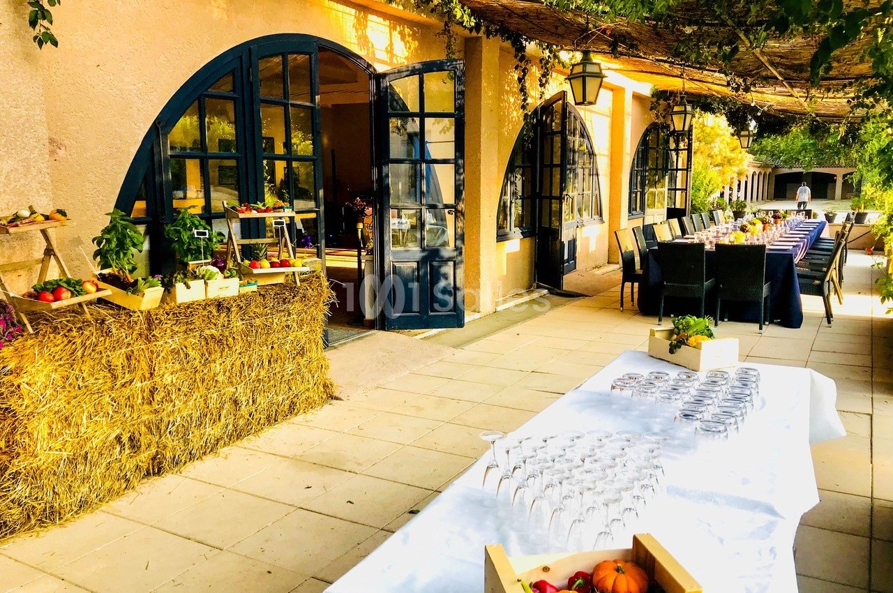 Terrasse ensoleillée avec tables dressées, verres alignés, étals de fruits et légumes, et décor végétal.