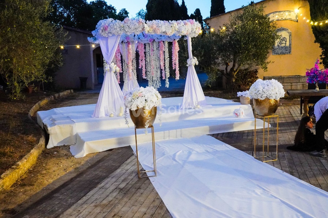 Allée blanche menant à une arche décorée de fleurs roses et blanches, entourée de pots fleuris et d'arbres.