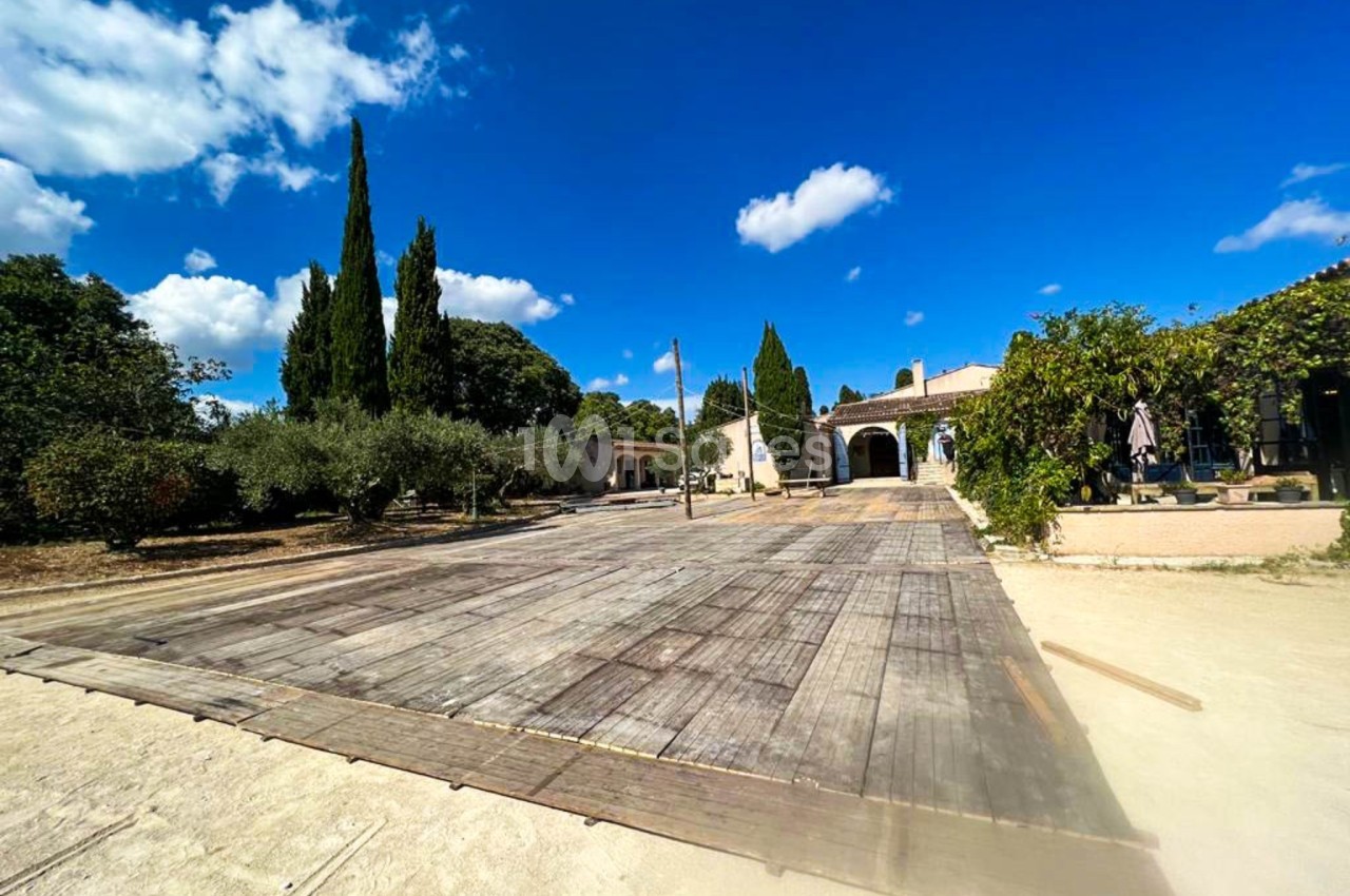 Cour extérieure ensoleillée avec une grande terrasse en bois, entourée d'arbres et d'un bâtiment de style méditerranéen.