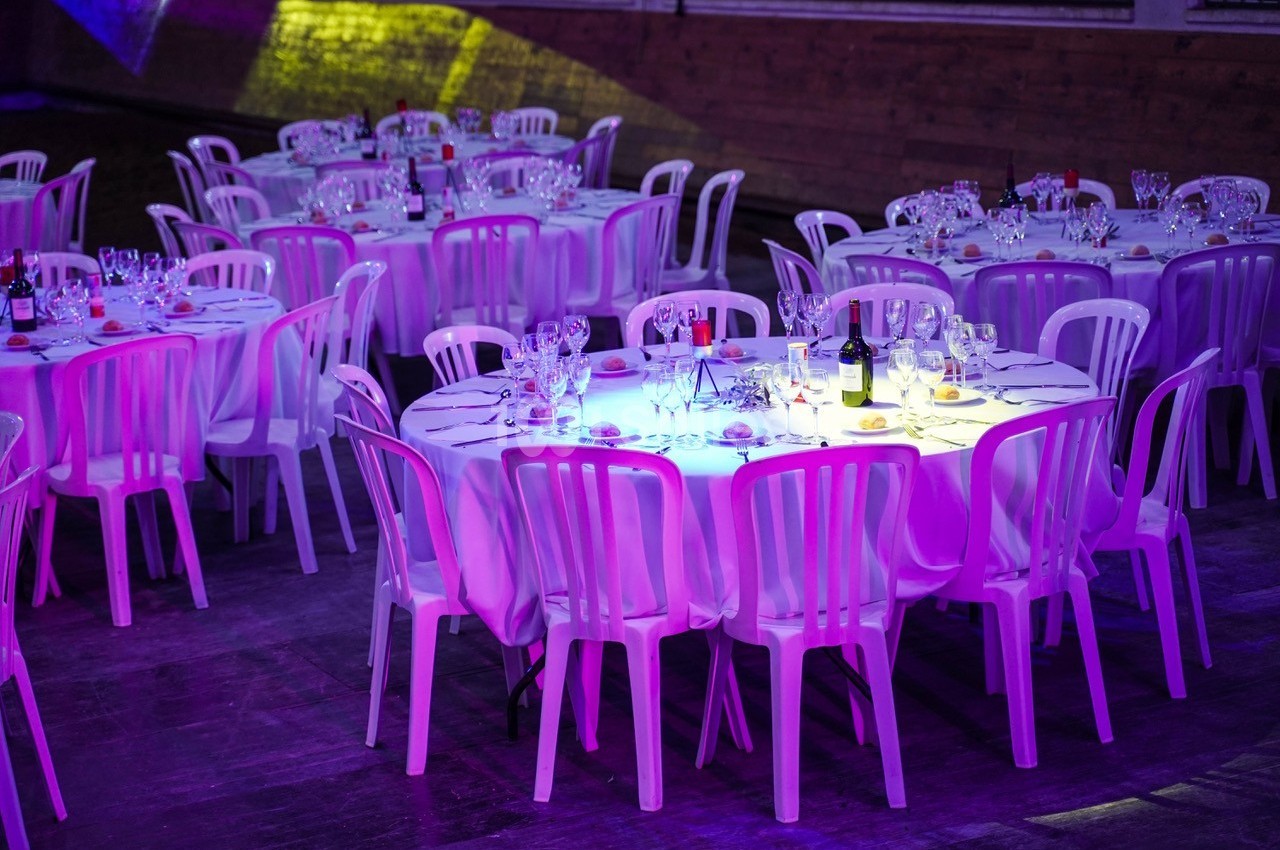 Tables rondes dressées avec nappes blanches, verres et bouteilles, entourées de chaises en plastique dans une salle éclairée.