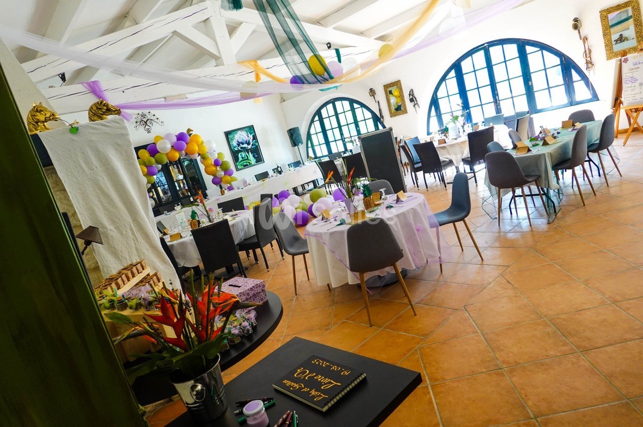 Salle décorée pour un événement avec tables dressées, ballons colorés et lumière naturelle entrant par de grandes fenêtres.