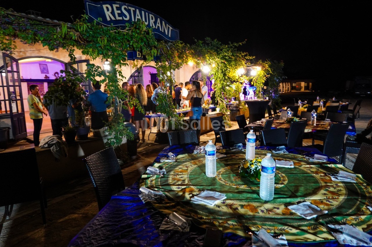 Tables dressées en extérieur avec nappes colorées, bouteilles d'eau et serviettes, devant un restaurant éclairé la nuit.