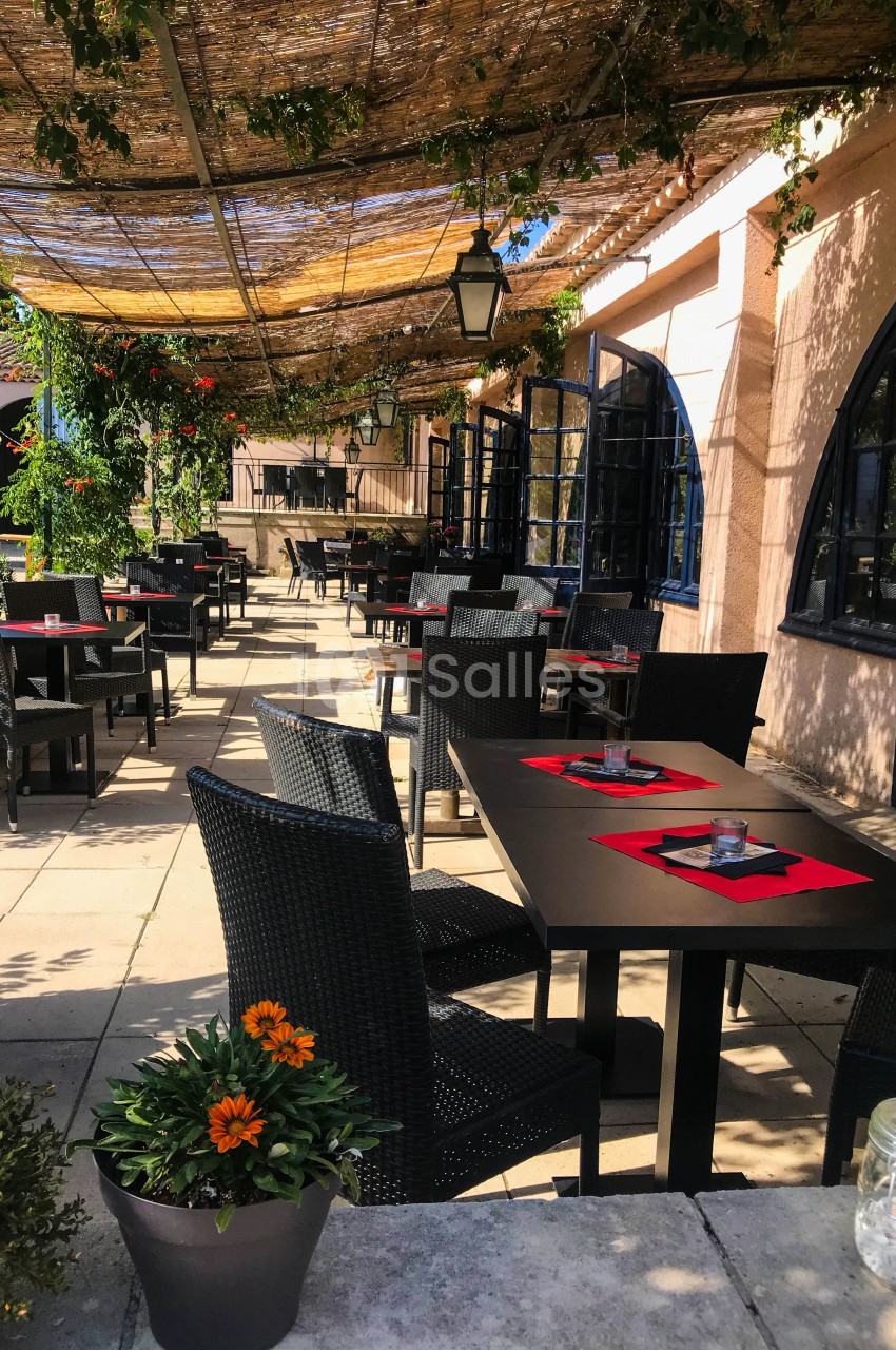 Terrasse extérieure d'un restaurant avec tables noires dressées, chaises en osier et pergola ombragée par des feuillages.