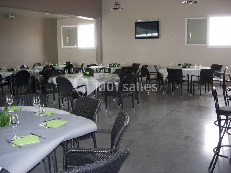 Salle de réception avec tables dressées, nappes blanches, serviettes vertes et chaises noires sur un sol en béton.