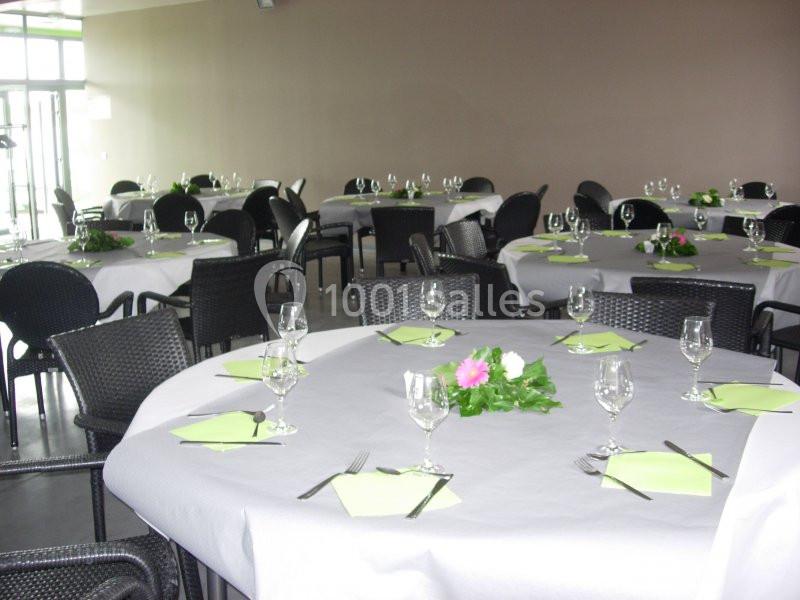 Salle avec des tables rondes dressées, nappes blanches, serviettes vertes, verres et décorations florales.