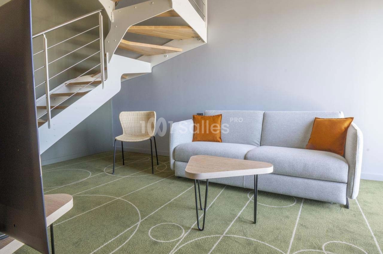 Salon moderne avec canapé gris, coussins orange, chaise en osier, table basse triangulaire et escalier en métal.