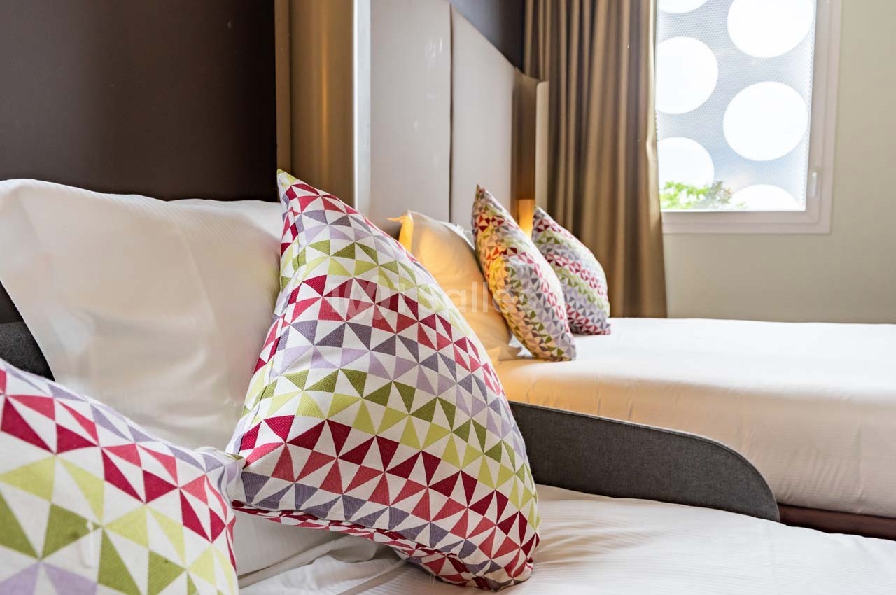Deux lits avec des coussins colorés à motifs géométriques dans une chambre lumineuse avec un rideau beige.