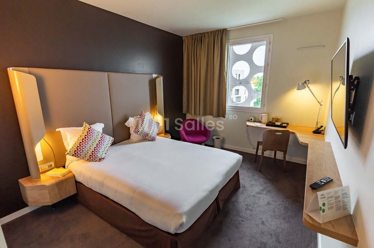 Chambre d'hôtel moderne avec lit double, bureau, fauteuil rose, télévision murale et fenêtre donnant sur l'extérieur.