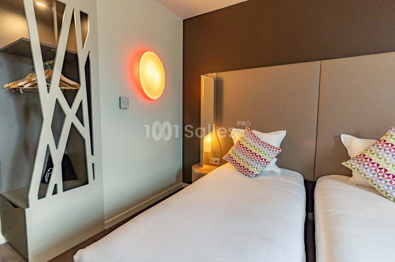 Deux lits simples avec têtes de lit rembourrées, coussins colorés, éclairage mural et espace de rangement ouvert.