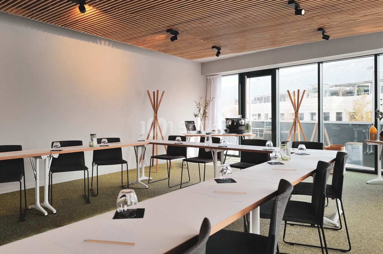 Salle de réunion lumineuse avec tables disposées en U, chaises noires, grandes fenêtres et éclairage au plafond.