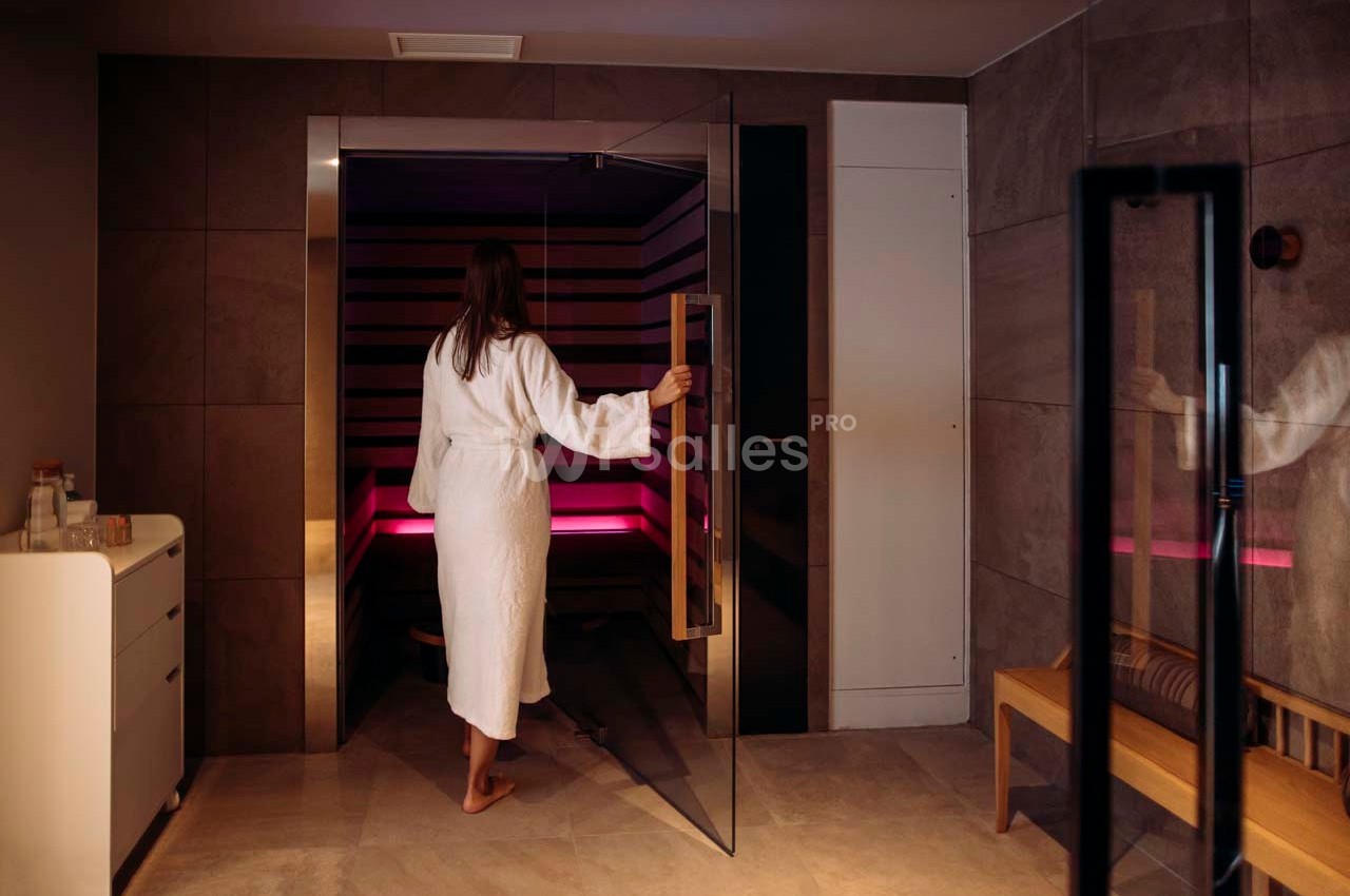 Une femme en peignoir entre dans un sauna moderne aux parois en bois, éclairé par une lumière tamisée.