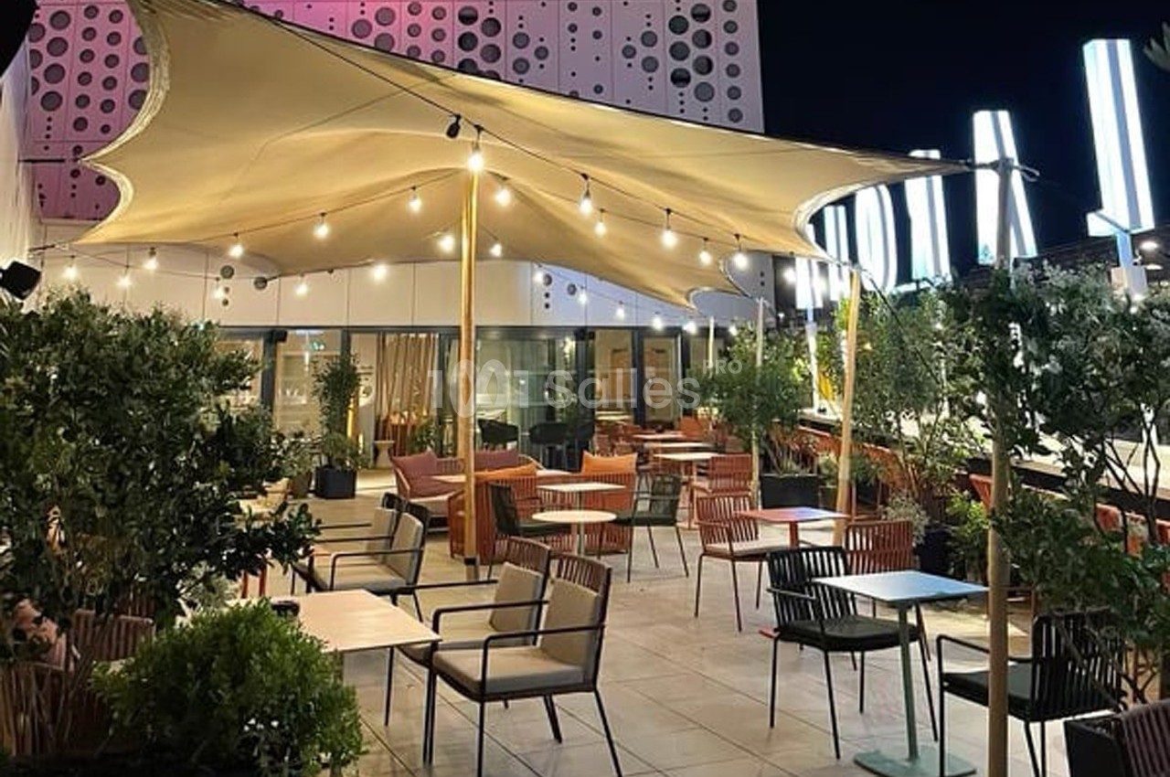 Terrasse extérieure éclairée de nuit avec tables, chaises, plantes et une toile tendue moderne en guise d'abri.