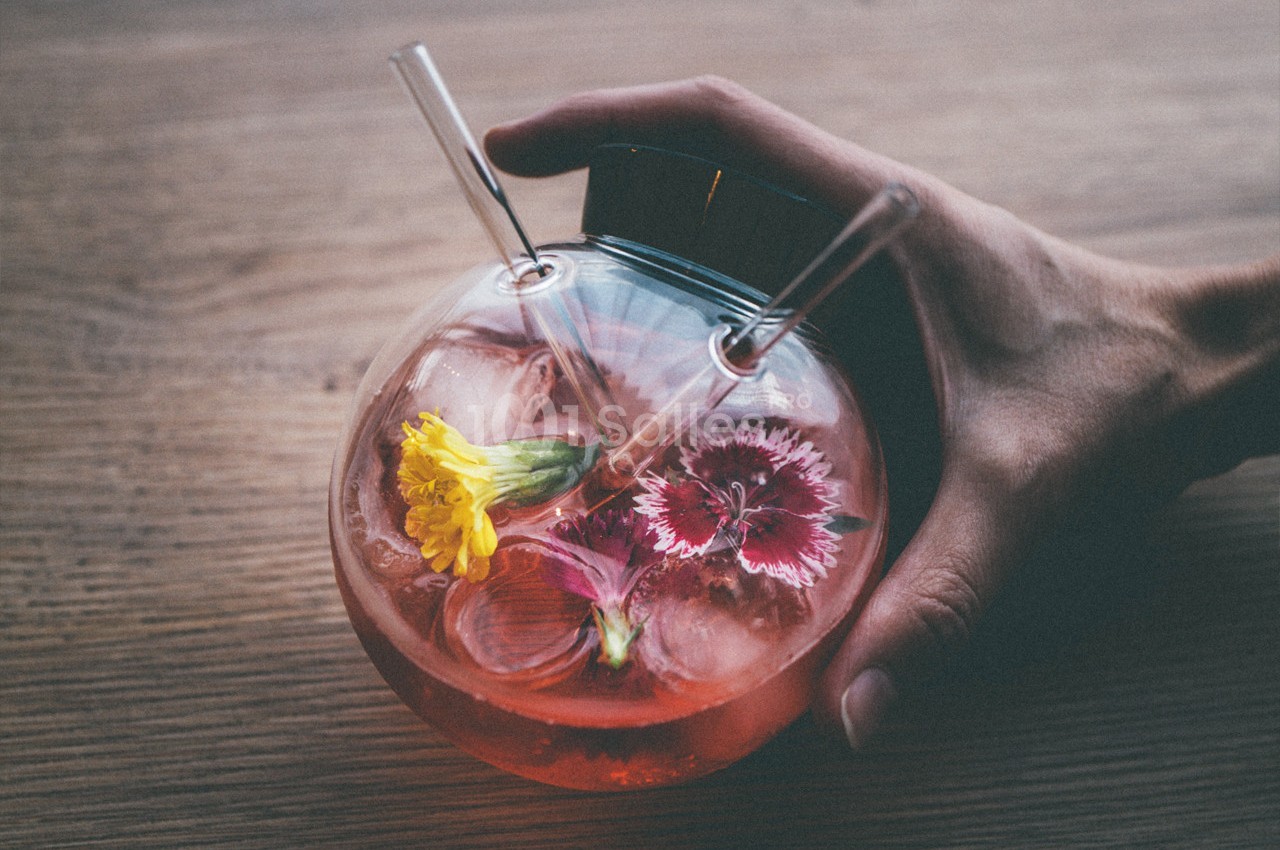 Main tenant un verre rond rempli d'une boisson rose, décorée de glaçons et de fleurs colorées, sur une table en bois.