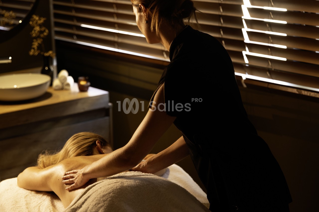 Une femme reçoit un massage relaxant dans une pièce tamisée avec des stores en bois et une ambiance apaisante.