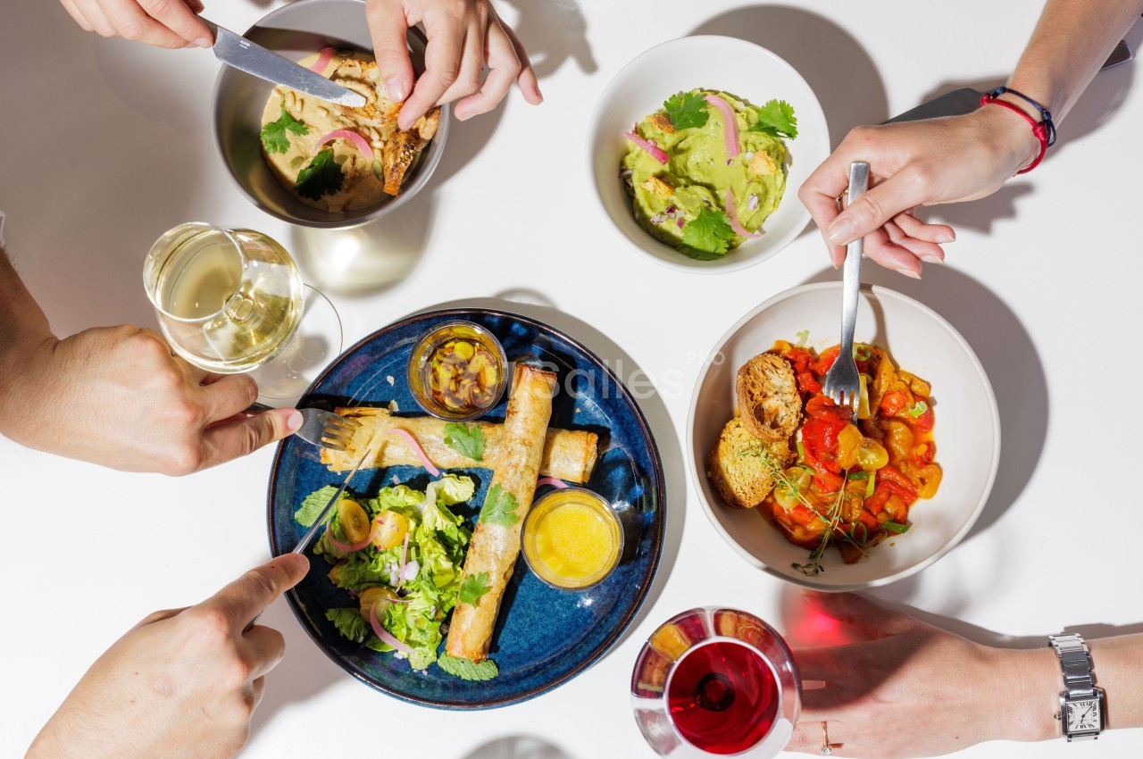 Repas convivial avec plusieurs plats colorés, des mains partageant des assiettes et des verres de vin sur une table blanche.