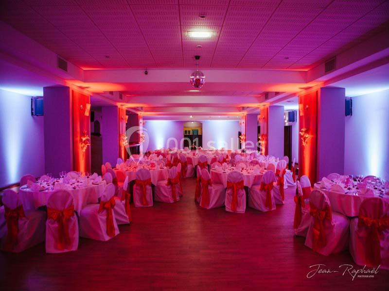 Salle de réception décorée avec des tables rondes, nappes blanches et chaises ornées de nœuds rouges, éclairage tamisé.