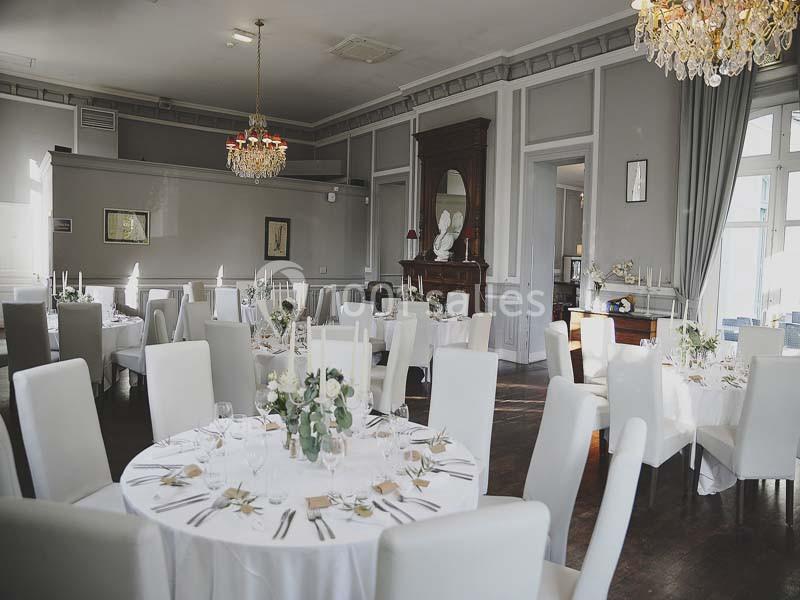 Salle de réception élégante avec tables rondes dressées, chaises blanches et lustres suspendus.