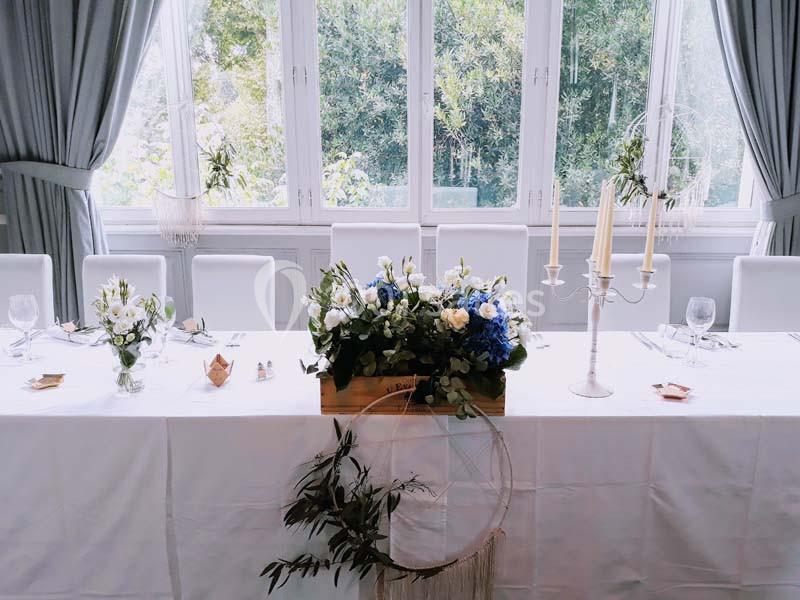 Table décorée pour un événement, ornée de fleurs blanches et bleues, bougies et éléments végétaux devant une grande fenêtre.