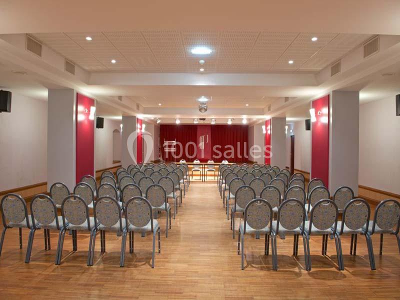 Salle de conférence avec rangées de chaises alignées face à une scène équipée d’un rideau rouge.