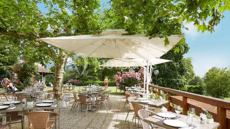 Terrasse ombragée avec tables et chaises en plein air, entourée de verdure et fleurs sous un ciel dégagé.