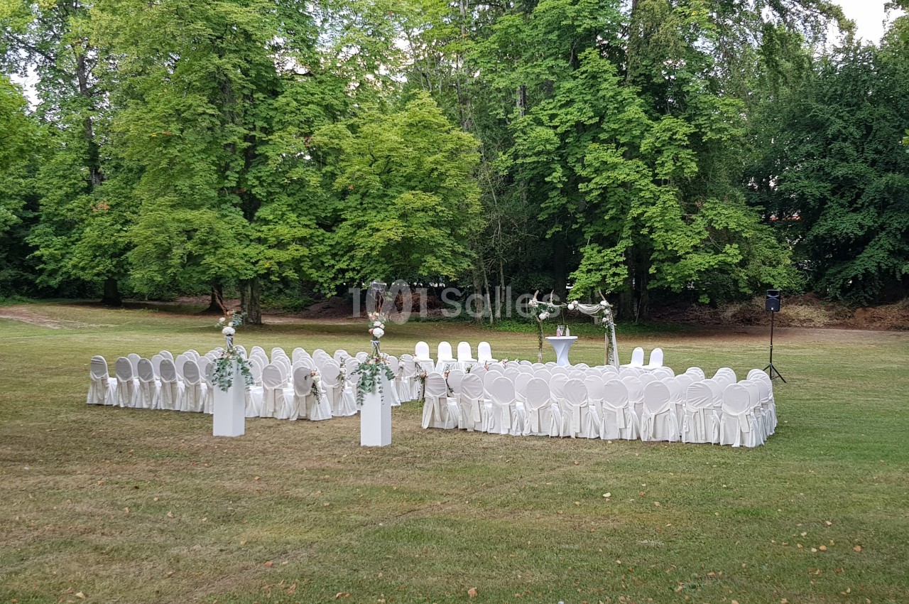 Chaises blanches disposées en cercle sur une pelouse, entourées d'arbres, avec une décoration florale et un pupitre central.