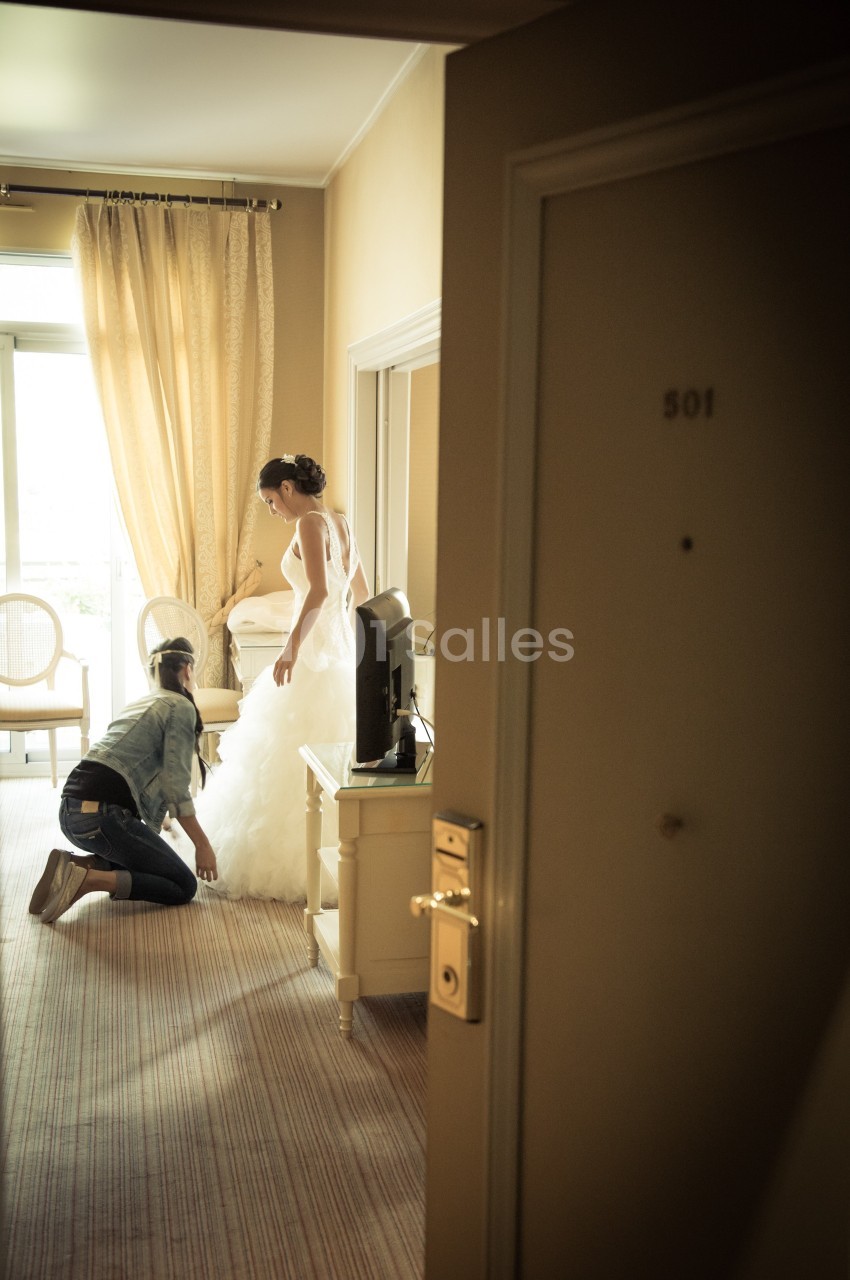 Une femme ajuste la robe de mariée d'une autre femme dans une chambre d'hôtel lumineuse.