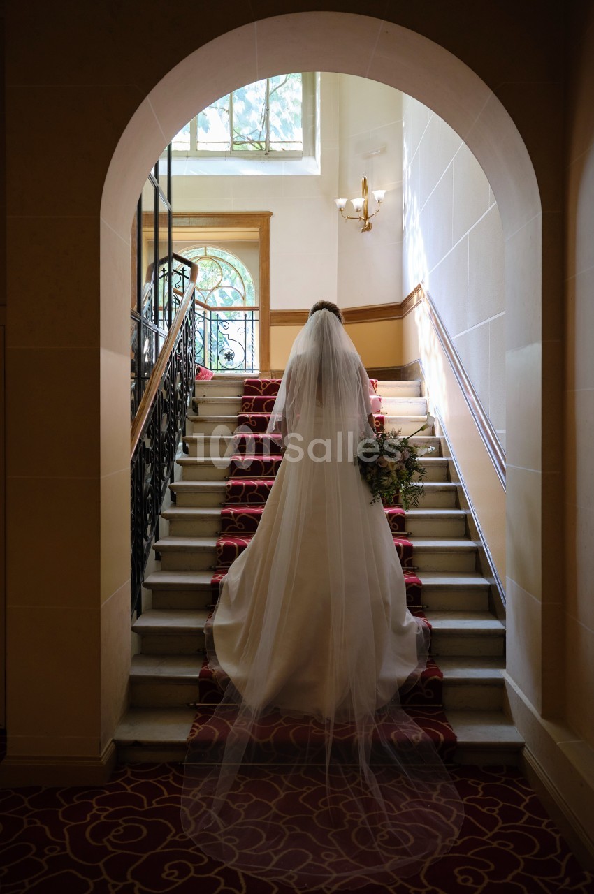 Une mariée en robe blanche monte un escalier décoré de tapis rouge dans un intérieur lumineux.