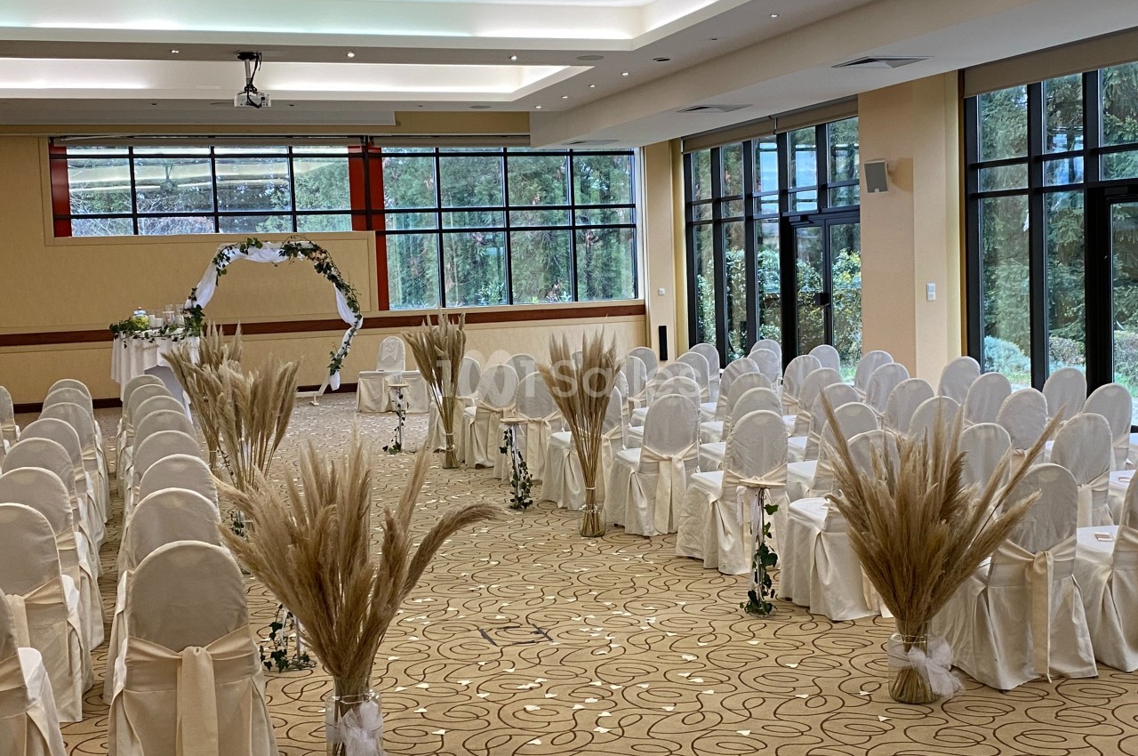 Salle de réception décorée pour une cérémonie, avec des rangées de chaises blanches et des ornements floraux.