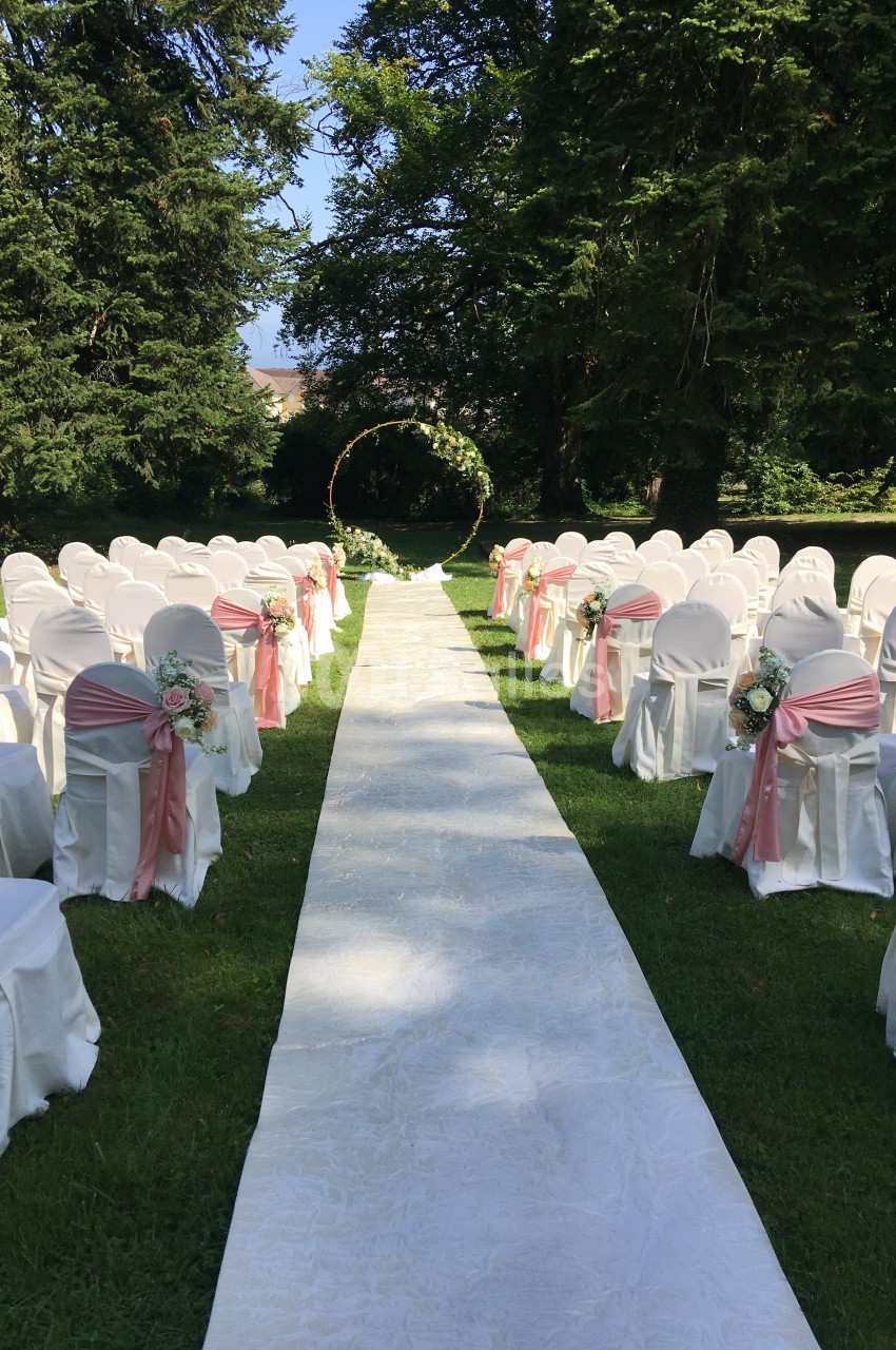 Allée centrale bordée de chaises blanches décorées de rubans roses, menant à une arche florale dans un jardin verdoyant.
