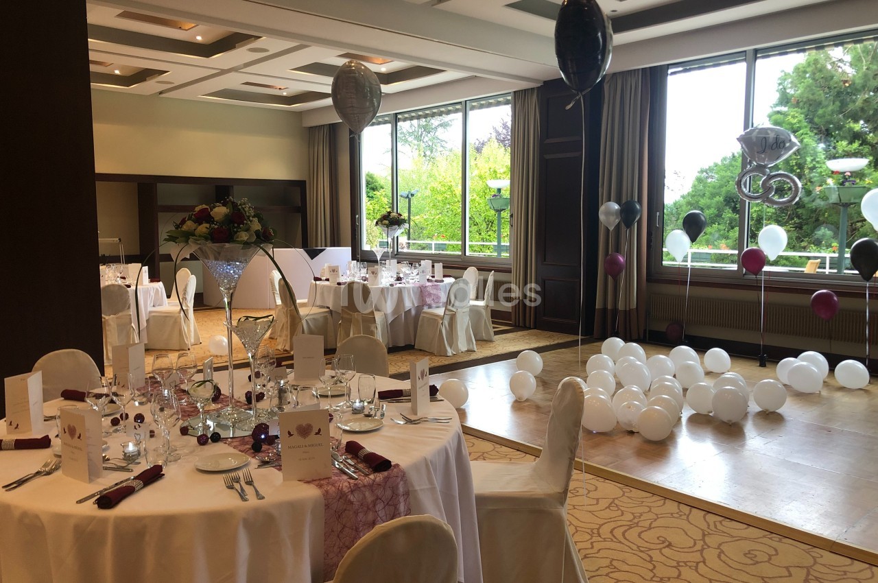 Salle décorée pour une réception avec tables dressées, ballons et grandes fenêtres donnant sur un jardin.