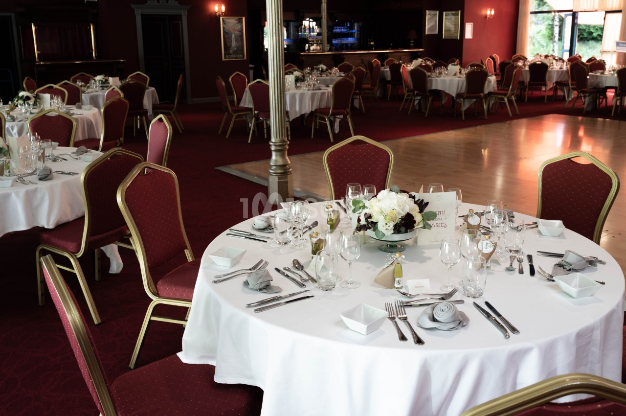 Salle de réception avec tables rondes dressées, nappes blanches, chaises rouges et piste de danse en bois.