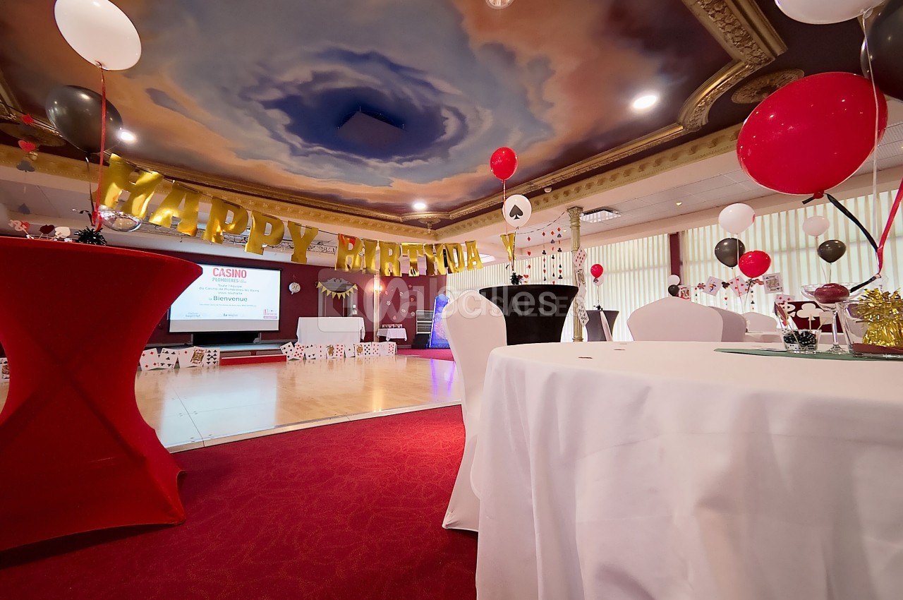 Salle décorée pour une fête, avec ballons rouges et noirs, tables dressées et une bannière ’Happy Birthday’.