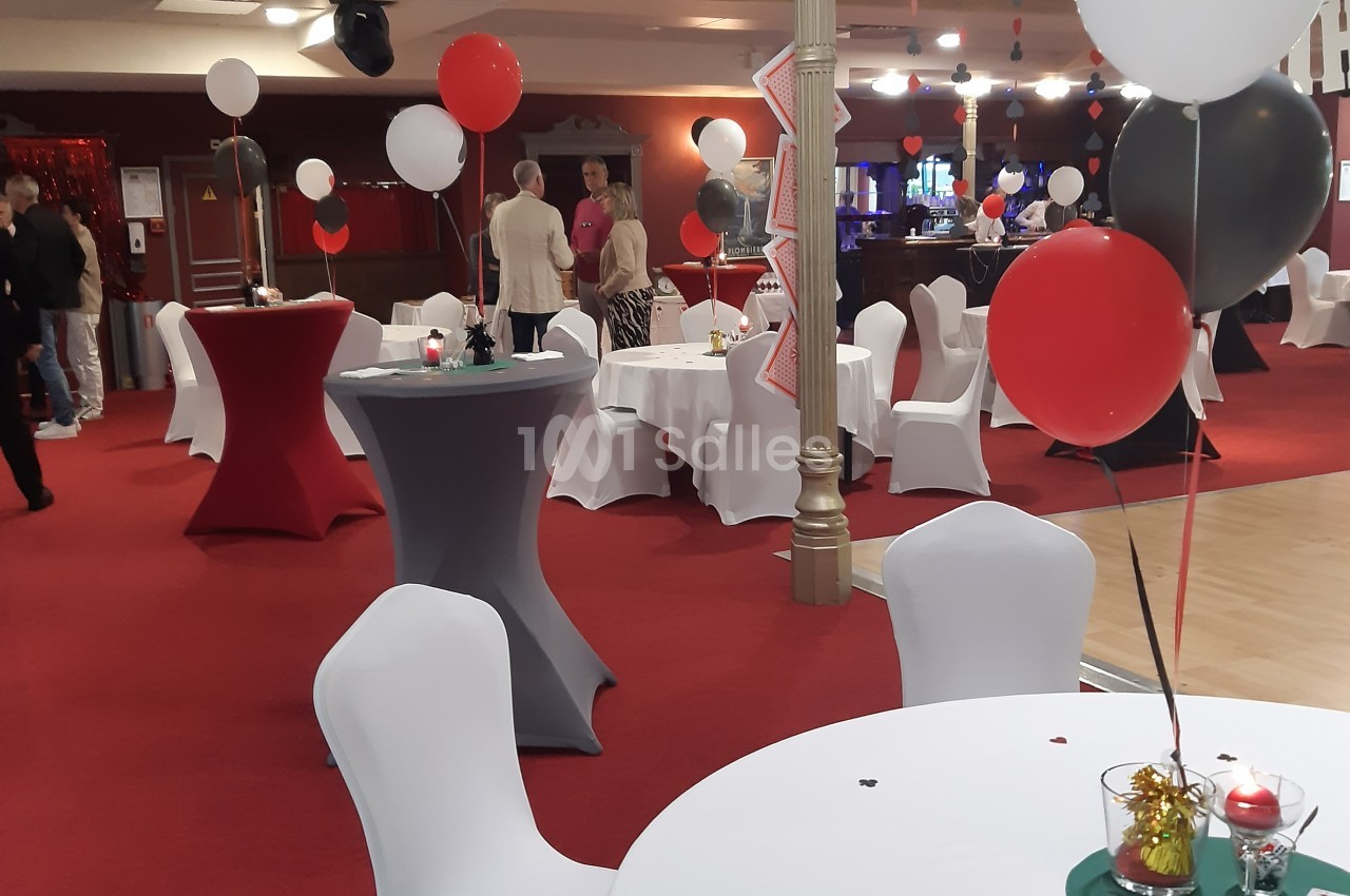 Salle décorée avec des ballons rouges, noirs et blancs, tables rondes couvertes de nappes blanches, ambiance festive.