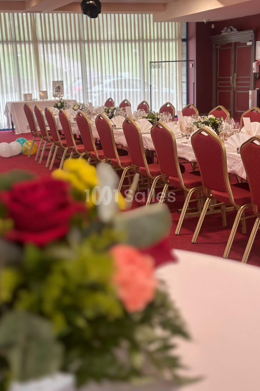 Salle de réception avec tables dressées, nappes blanches, chaises rouges et décorations florales au premier plan.