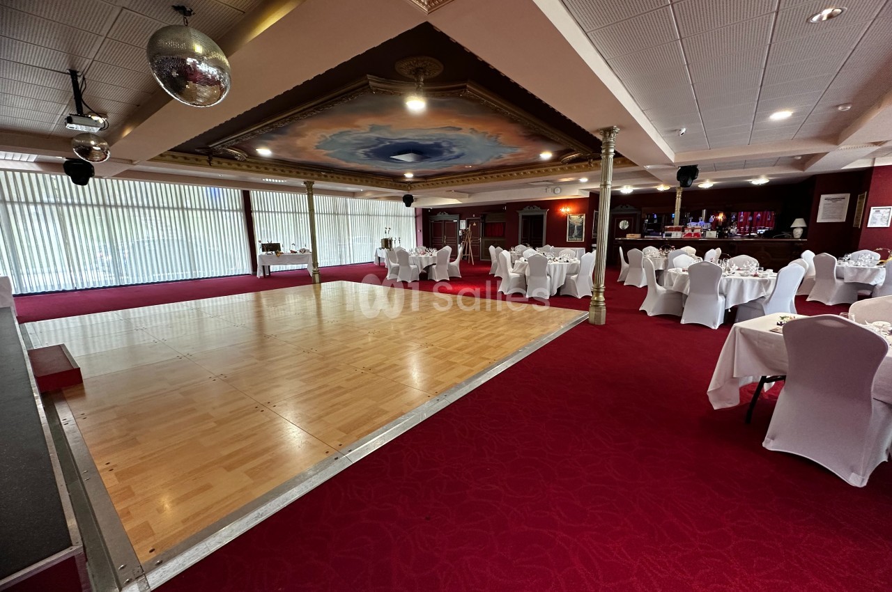 Salle de réception avec piste de danse en bois, tables dressées et chaises blanches sur une moquette rouge.
