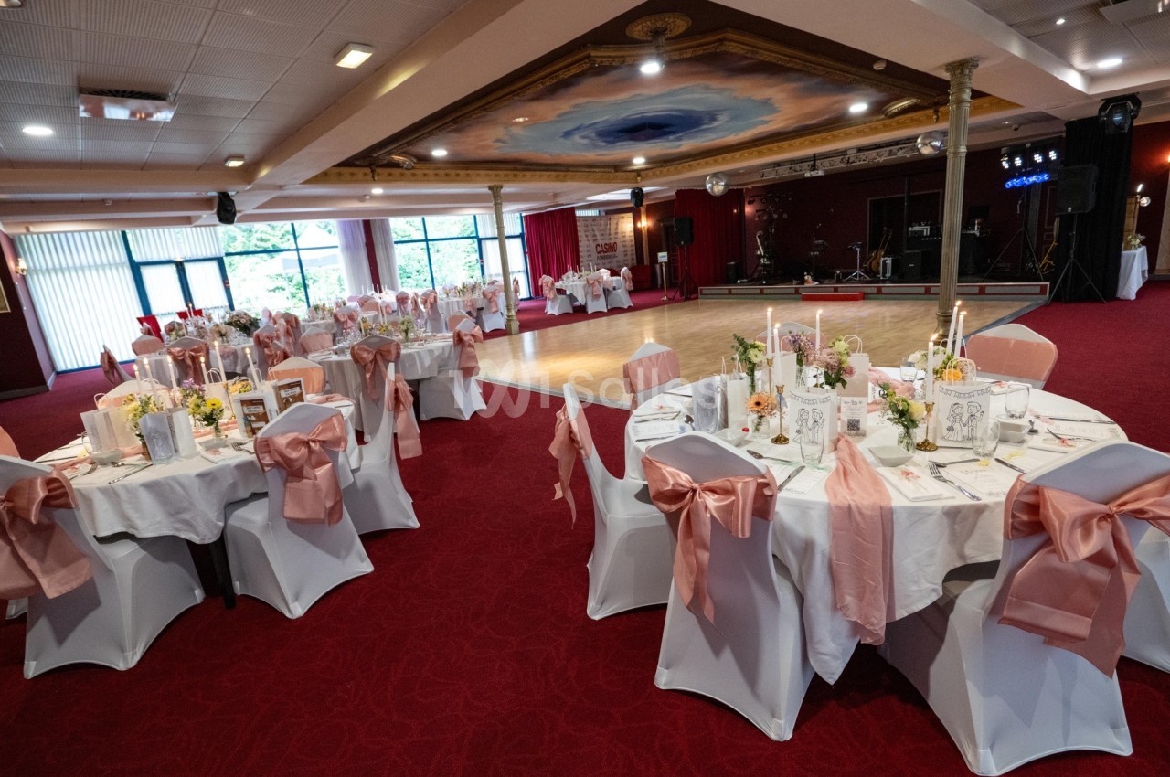 Salle de réception décorée avec des tables rondes, nappes blanches, chaises ornées de nœuds roses et bougies allumées.