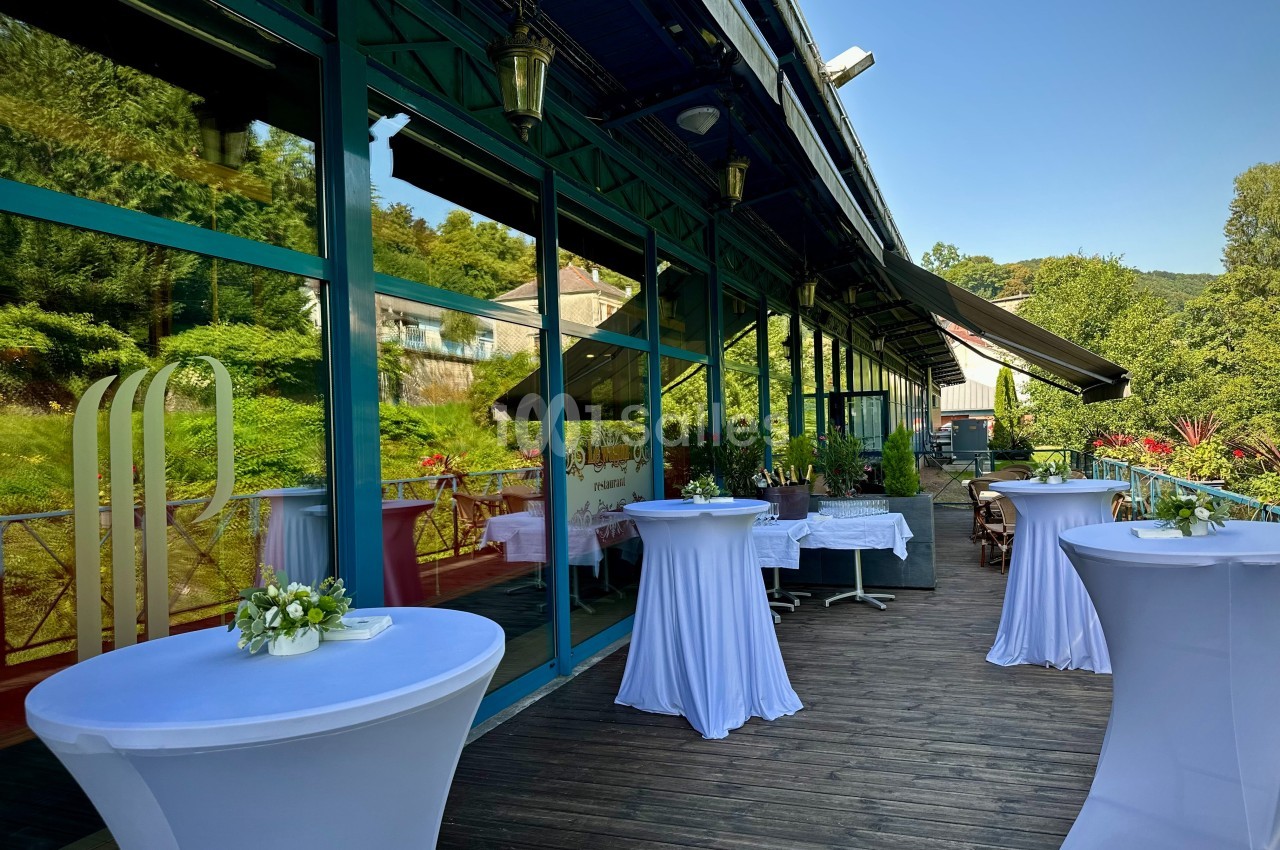 Terrasse en bois avec tables hautes drapées de blanc, bordée de grandes baies vitrées et entourée de verdure.