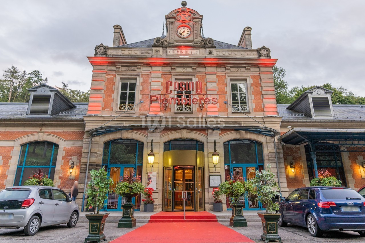 Façade d'un bâtiment historique marqué ’Casino Plombières’, avec un tapis rouge et des voitures stationnées.
