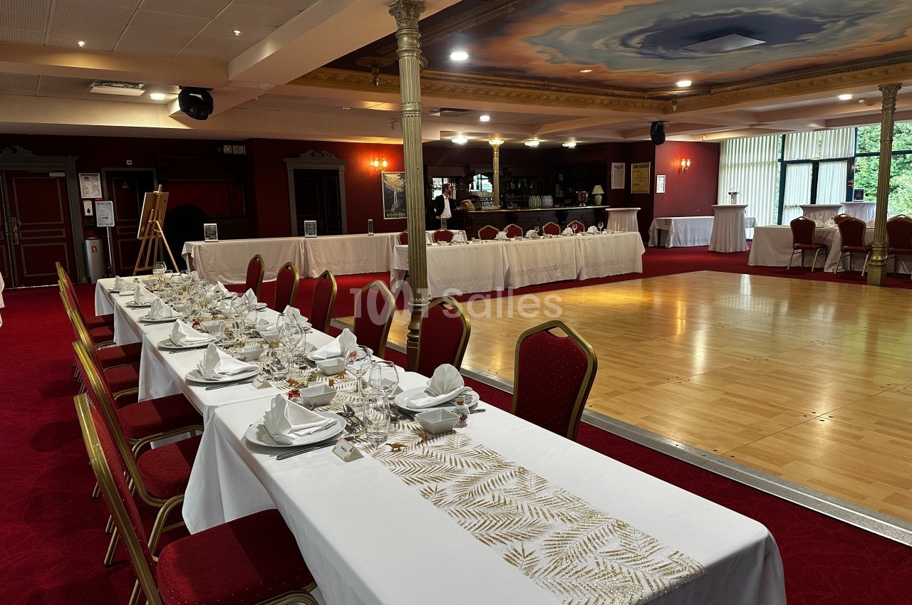 Salle de réception avec tables dressées, chaises rouges, piste de danse en bois et bar en arrière-plan.