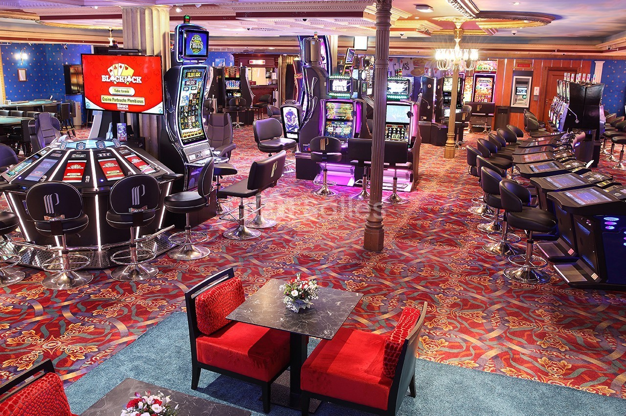 Salle de casino avec machines à sous, tables de jeux électroniques et sièges rouges sur un sol à motifs colorés.